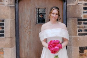 Bride in off-shoulder white gown holding vibrant pink peony bouquet, standing before rustic wooden door.