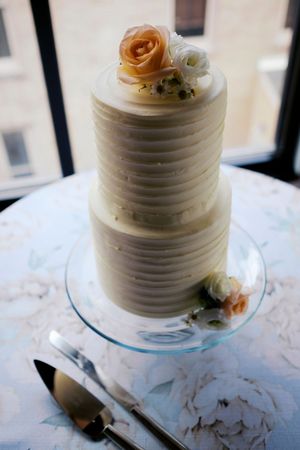 Elegant two-tier white wedding cake with textured frosting and peach roses on a glass stand near a window.