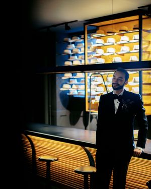 Man in tuxedo standing in dimly lit bar with illuminated hat display shelves in background.