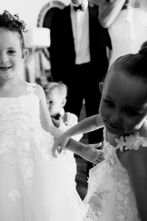 Two young flower girls in white lace dresses smile and interact at a wedding ceremony in black and white photo.