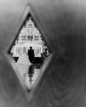 Black and white wedding photo through diamond-shaped opening showing bride and groom at ornate altar.
