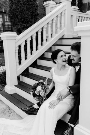 Black and white photo of a laughing bride and groom sitting on outdoor staircase steps, bride holding a bouquet.
