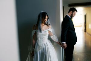 Bride in off-shoulder wedding gown with veil holds groom's hand in suit in a softly lit hallway.