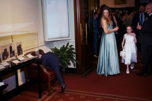 Woman in teal gown and flower girl enter elegant hall as man bends over desk at formal wedding event.