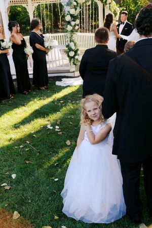 A young flower girl in a white tulle dress smiles at an outdoor wedding ceremony with bridesmaids in black gowns.