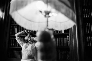 Black and white photo of a bride in embroidered lehenga adjusting her hair near a lamp in an elegant library.