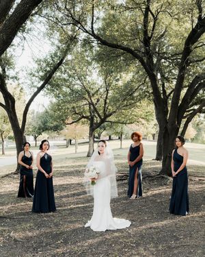 Bride in white gown holds bouquet surrounded by four bridesmaids in navy dresses under trees in a park setting.