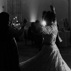 Black and white photo of two women dancing at a wedding reception in elegant embroidered gowns.