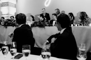 Black and white wedding reception photo of guests seated at tables watching a speech at the head table.