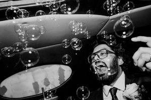 Black and white wedding photo of groom laughing among floating bubbles at reception celebration.