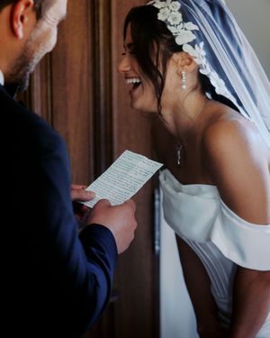 Bride in white off-shoulder gown laughs joyfully as groom reads wedding vows from paper during ceremony.