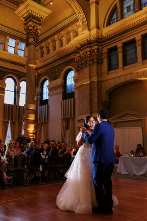 Bride and groom share first dance in elegant gold ballroom with arched windows and guests seated around the dance floor.