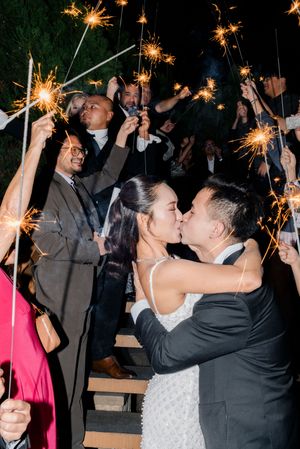A newlywed couple shares a kiss during a romantic sparkler send-off surrounded by cheering wedding guests at night.