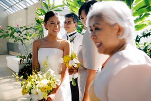 Bride in white strapless gown holds yellow floral bouquet, smiling at guests during an elegant indoor wedding ceremony.