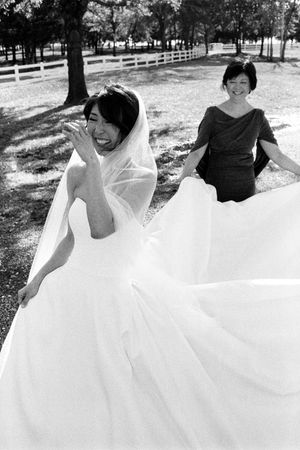 Black and white photo of a bride in a flowing white gown and veil, laughing outdoors while a woman helps arrange her dress.