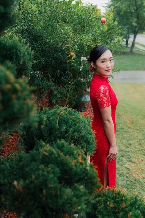 Woman in red lace cheongsam qipao dress posing gracefully among lush green garden shrubs and trees.