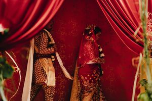 Two individuals in ornate red and gold Indian bridal attire stand before rich crimson drapes at a traditional wedding ceremony.