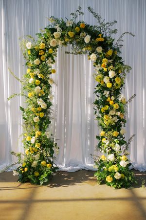 Elegant wedding arch adorned with yellow and white roses, lush greenery, set against flowing white drape backdrop.