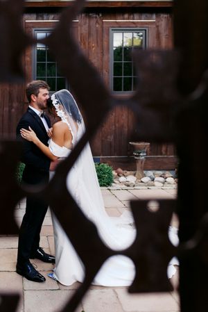 Bride and groom share a romantic embrace outside a rustic barn venue, framed by ornate iron gates.