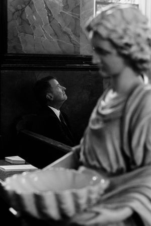 Black and white photo of a man in suit gazing upward in a church pew beside a classical stone angel statue.