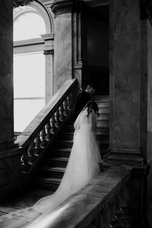 Black and white photo of a bride in a flowing gown and veil ascending a grand staircase in an elegant venue.