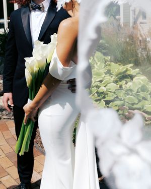 Bride in white gown holding calla lily bouquet stands beside groom in black tuxedo outdoors at wedding venue.
