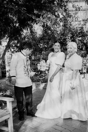 Black and white photo of a bride in a white gown with groom and elderly woman at an outdoor wedding ceremony.