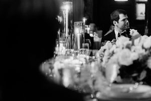 Black and white wedding reception photo with groom in tuxedo seated at elegantly decorated table with candles and flowers.