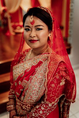 A bride in a red embroidered saree with gold jewelry and red veil poses elegantly for her traditional Indian wedding.