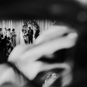 Black and white wedding ceremony photo captured through blurred foreground, showing couple at floral arch altar.