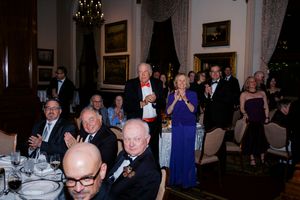 Elegantly dressed guests applaud at a formal dinner event in an ornate dining room with chandeliers and artwork.