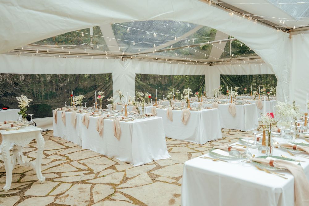 Elegant outdoor wedding reception tent with white draped tables, floral centerpieces, and string lights on stone flooring.