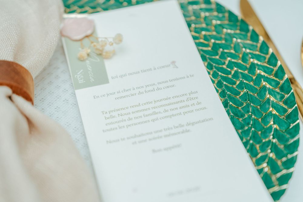 Elegant wedding menu card on teal woven placemat with gold accents and pink floral decoration.