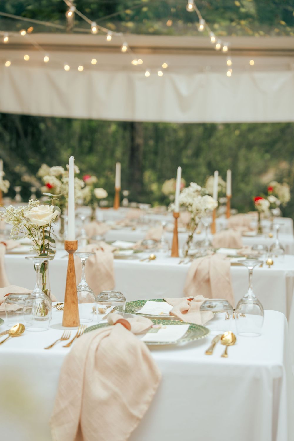 Elegant outdoor wedding reception tables with white linens, gold candlesticks, blush napkins, and floral centerpieces under string lights.