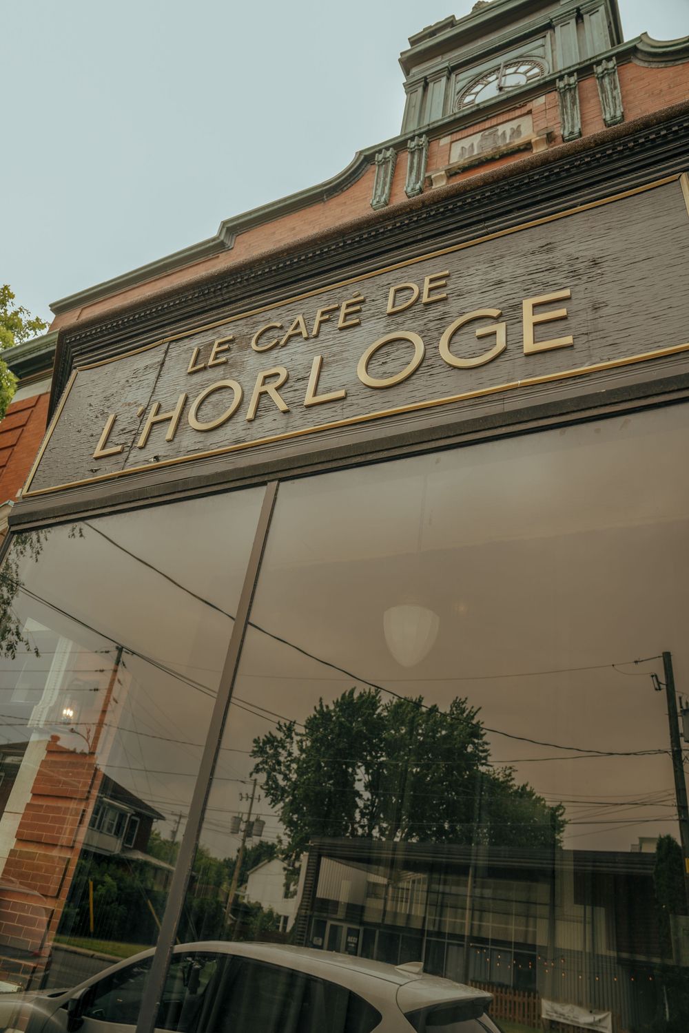 Exterior view of Le Cafe de L'Horloge with gold lettering on a dark facade and brick building above.