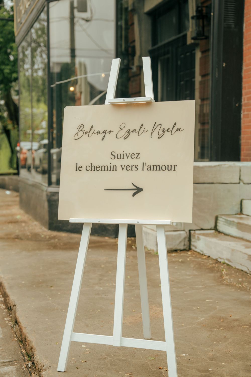 White easel sign reading Suivez le chemin vers l'amour with arrow, wedding directional sign on sidewalk.