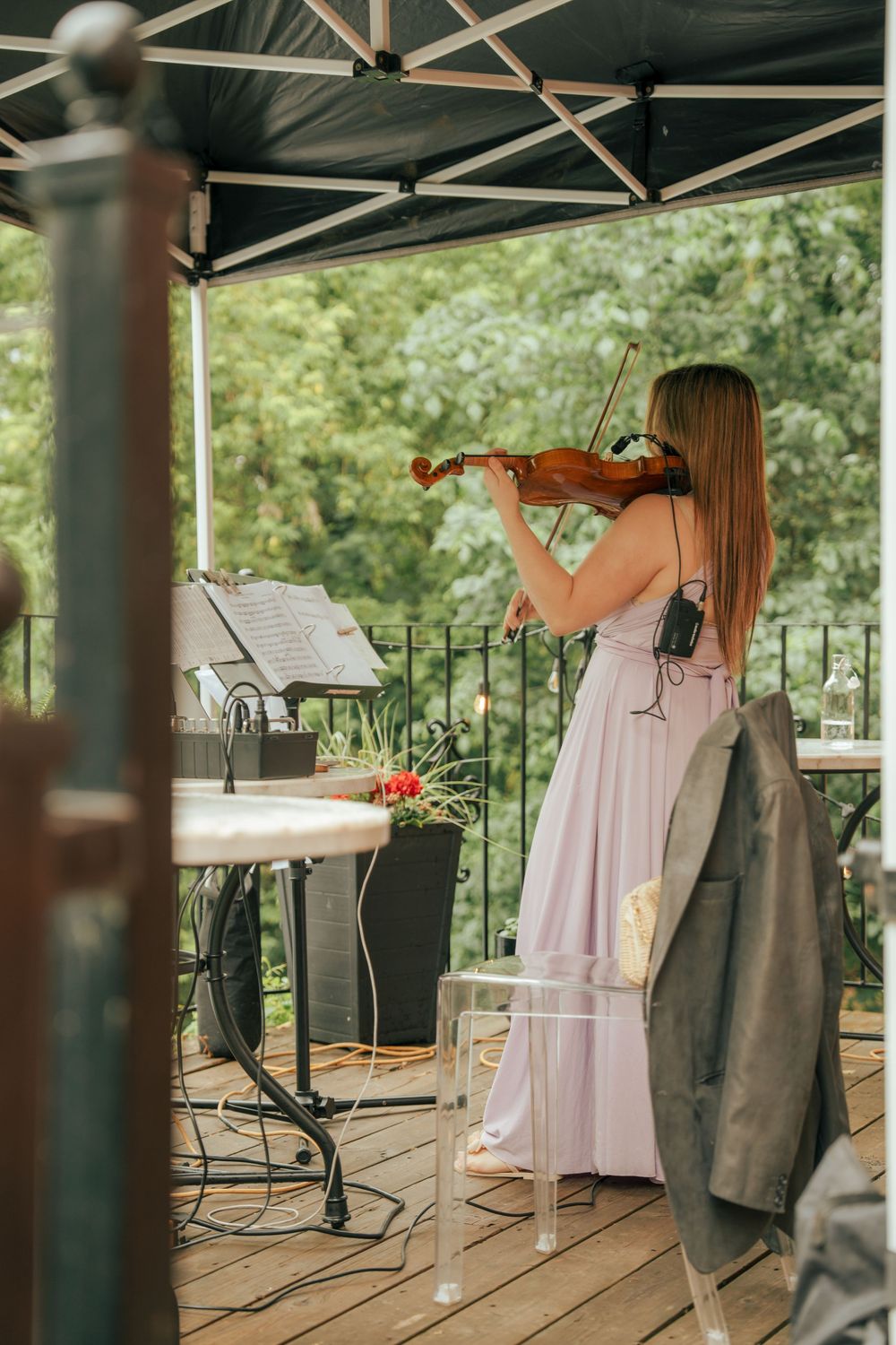 A woman in a pink gown plays violin on an outdoor wooden deck stage under a black canopy tent.