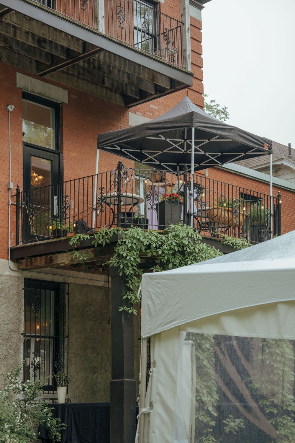 Brick building exterior with wrought iron balcony, black canopy umbrella, lush greenery, and white event tent.