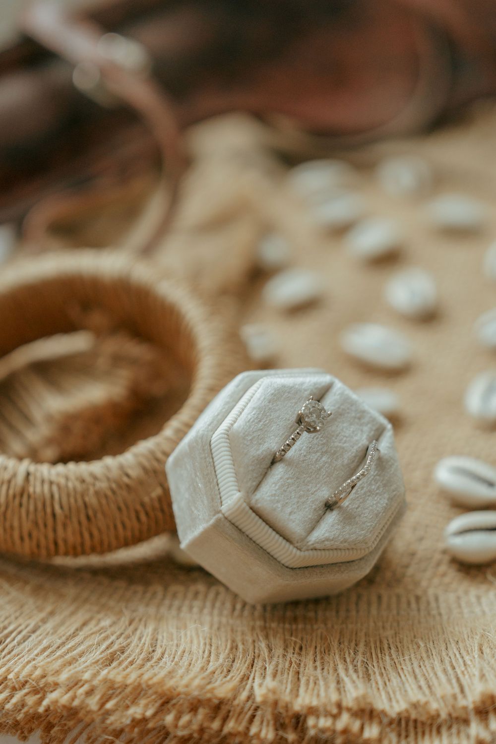 Elegant octagonal velvet ring box with delicate jewelry displayed on burlap fabric surrounded by cowrie shells and woven bracelet.