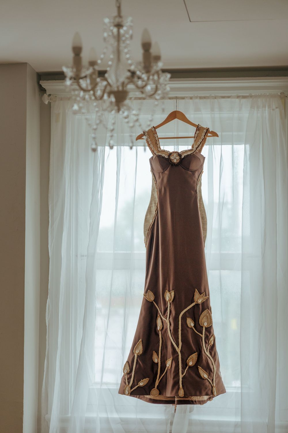 Elegant brown mermaid gown with gold floral appliques hanging on a wooden hanger in front of sheer white curtains.