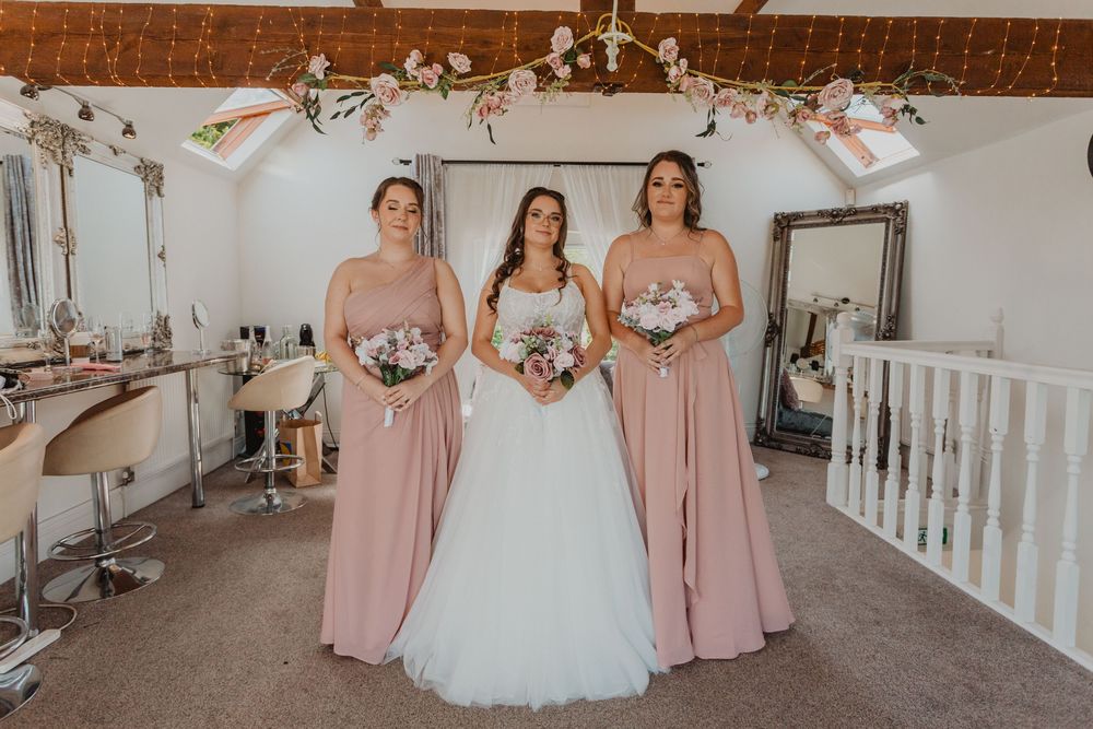 Bride in white gown flanked by two bridesmaids in blush pink dresses holding bouquets in an elegant bridal suite.