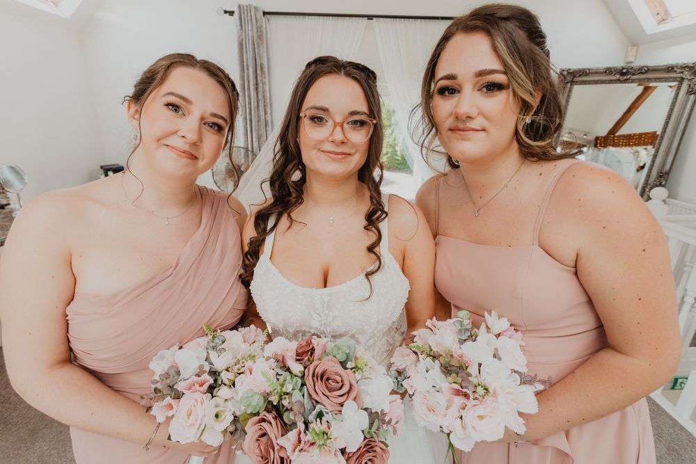 Bride in white gown holds pink bouquet flanked by two bridesmaids in blush pink dresses at wedding.
