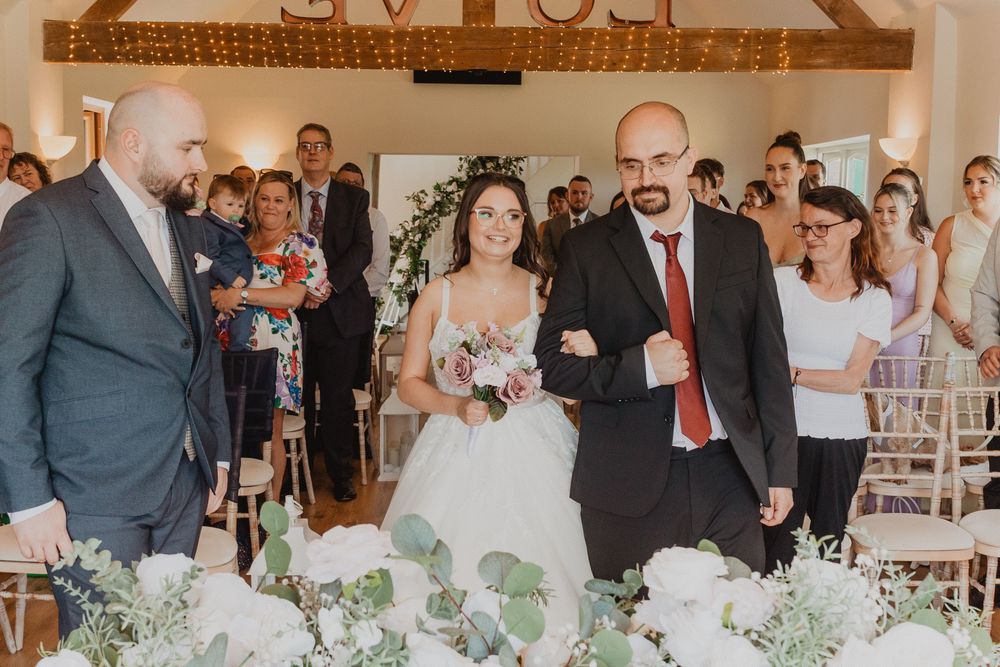 Bride walks down the aisle with escort at elegant wedding ceremony, guests watching, floral arrangements in foreground.