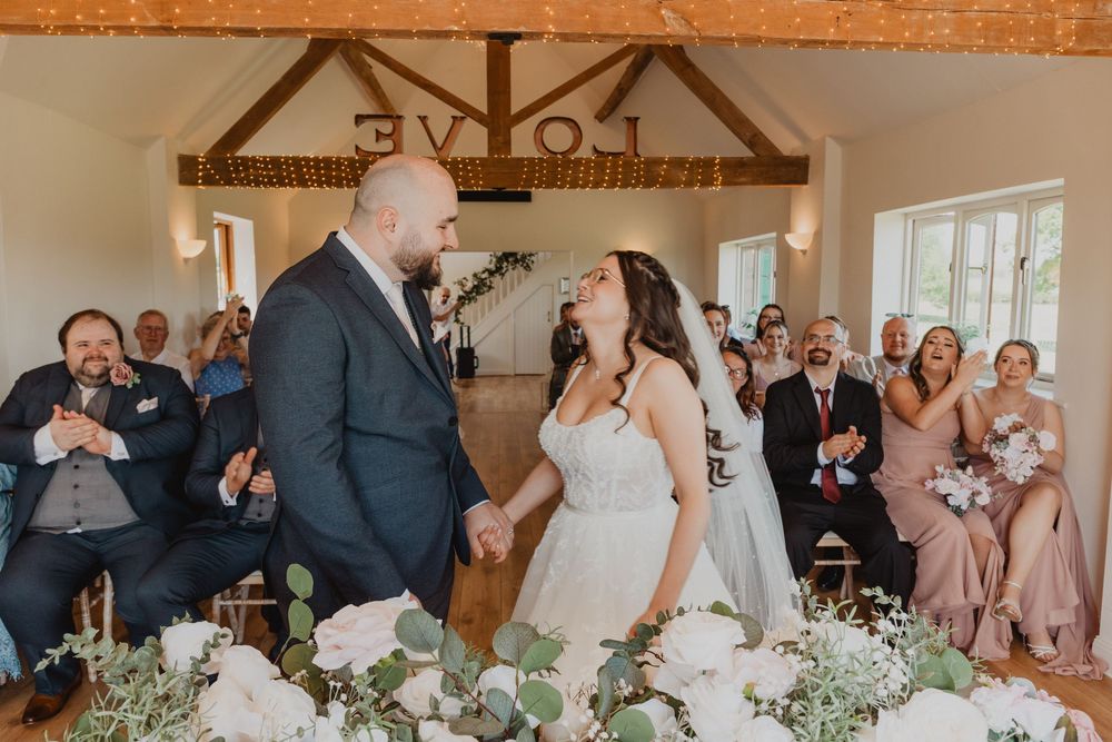 Joyful newlyweds hold hands and laugh during their wedding ceremony in a rustic barn venue with guests applauding.