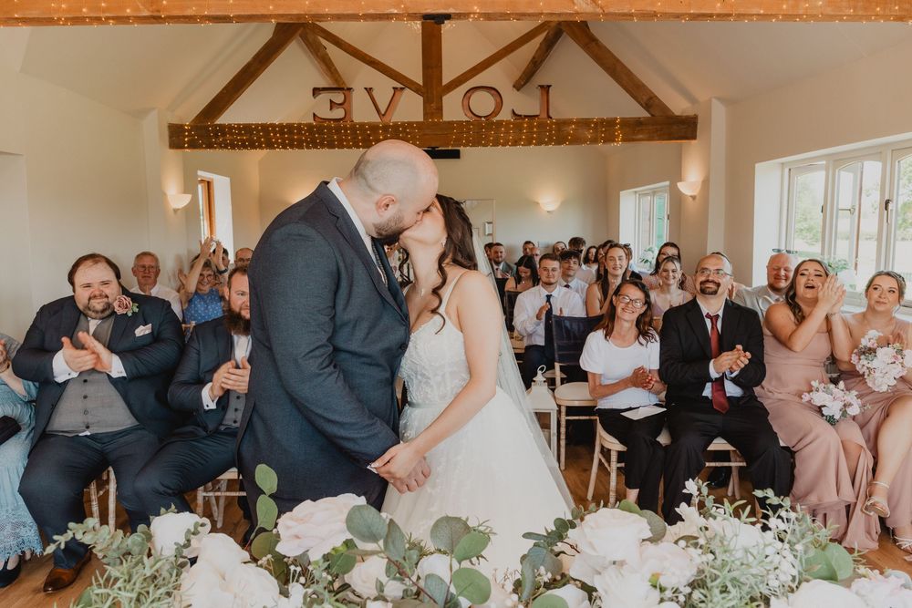 Bride and groom share first kiss at wedding ceremony while guests cheer in a rustic barn venue decorated with white flowers.