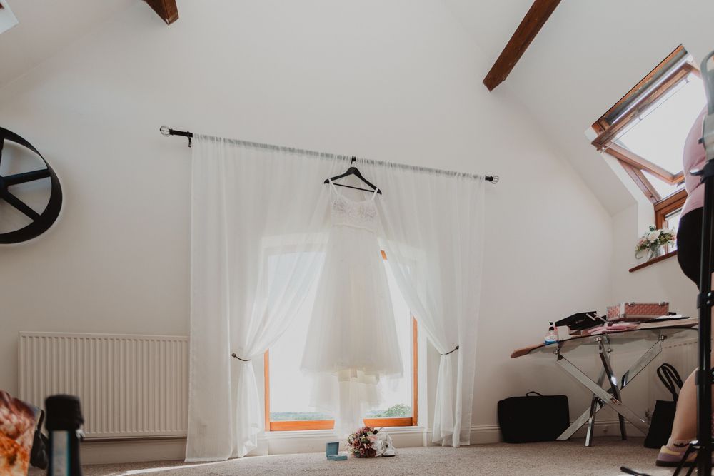 A white wedding dress hanging on a hanger in front of a bright window in a white attic room with wooden beams.