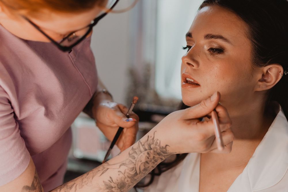 A tattooed makeup artist carefully applies makeup to a young woman's face in a bright studio setting.