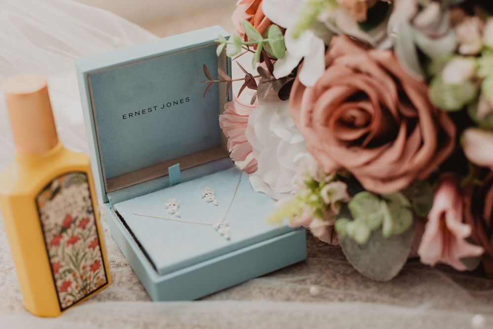 Ernest Jones teal jewelry box with delicate necklace, beside pink roses bouquet and Gucci perfume bottle.