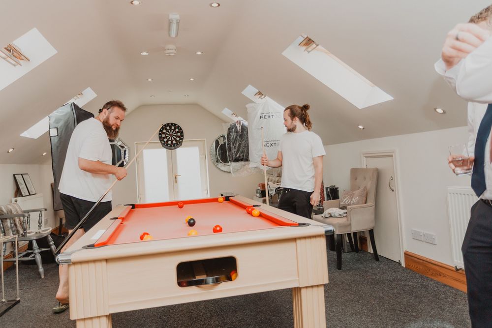 Two men playing pool on an orange-felt billiard table in a bright attic games room with skylights and dartboard.