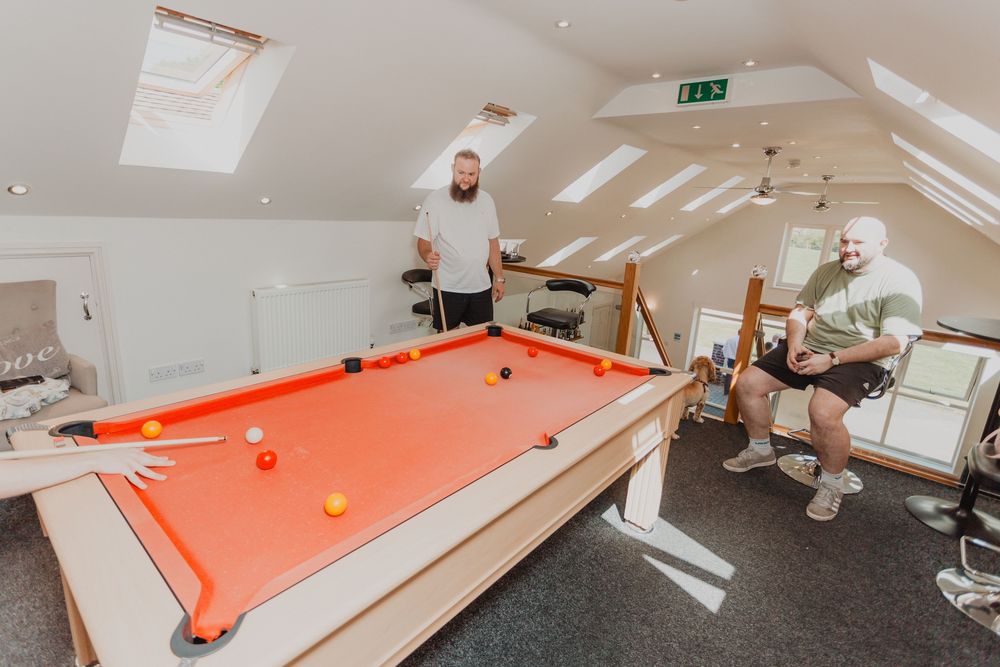 Two men enjoy a game of pool on an orange billiard table in a bright attic games room with skylights.
